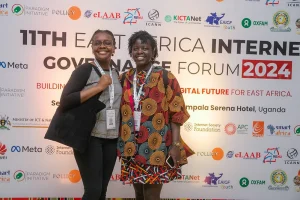 Nzambi J.Kakusu, Program Administrator, KICTANet, and Mitchelle Atieno standing behid the EAIGF 2024 banner during the East Africa Internet Governance Forum 2024.